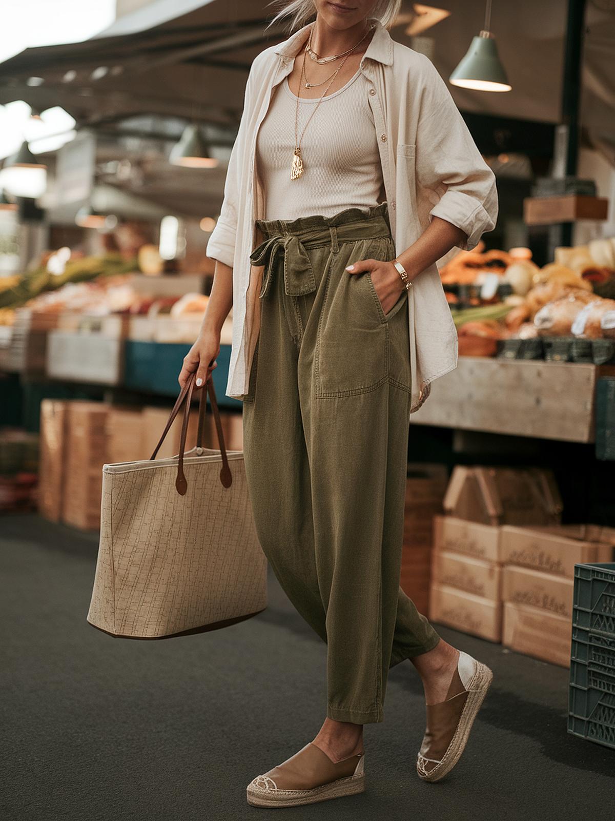 Woman in casual outfit shopping at outdoor market, carrying a beige tote bag and wearing green pants and slip-on shoes.