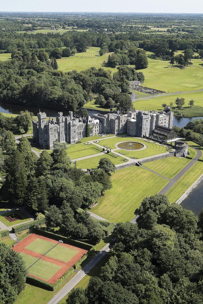Aerial view of an elegant castle surrounded by lush greenery, gardens, and tennis courts on a sunny day.