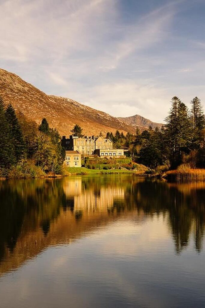 Elegant lakeside castle with mountain backdrop and lush greenery under a clear blue sky. Scenic reflection in water.