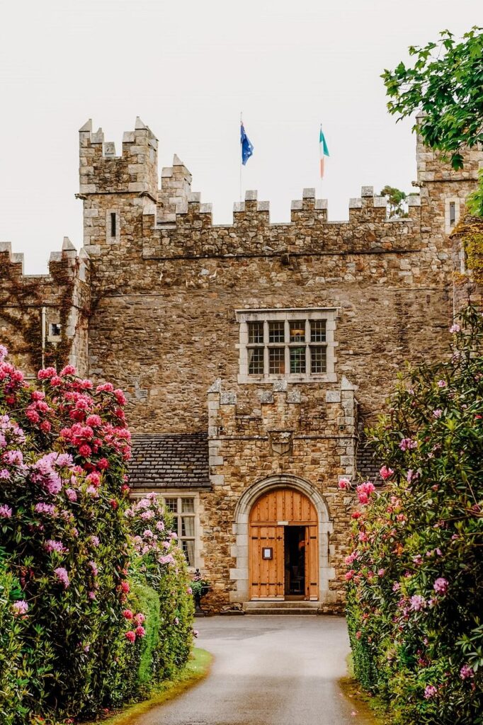 Historic stone castle entrance with blooming pink flowers and flags flying above under a cloudy sky.