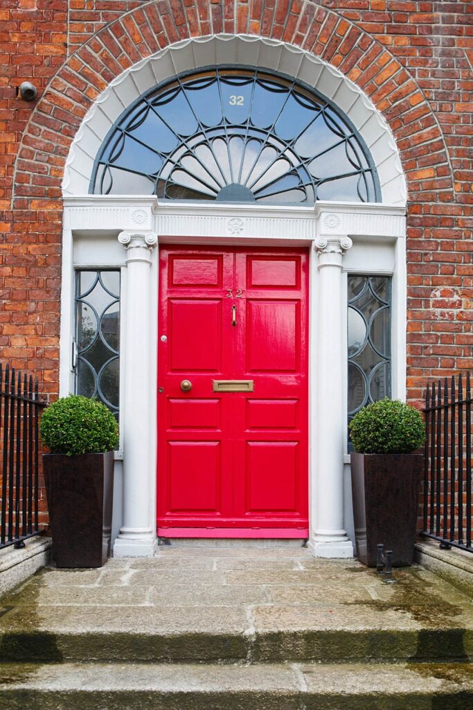 Red door with arch window and brick facade, flanked by columns and plants in pots. Elegant entrance design.