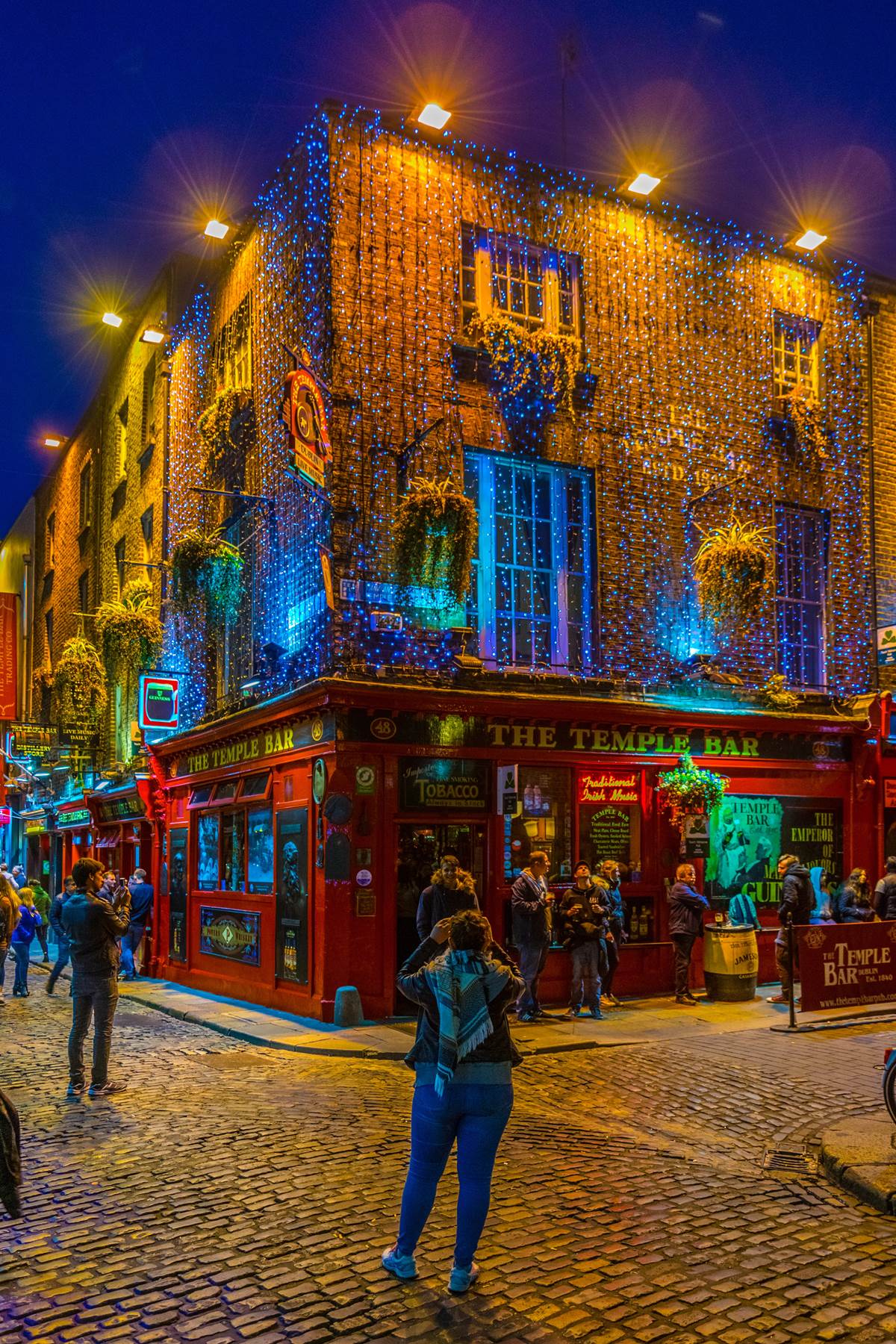 Colorful lights on historic bar facade at night with people taking photos, lively street scene.