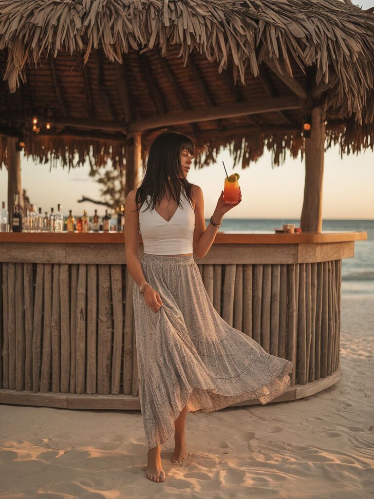 Person enjoying a tropical cocktail at a beach bar during sunset, wearing a flowy skirt and white top.