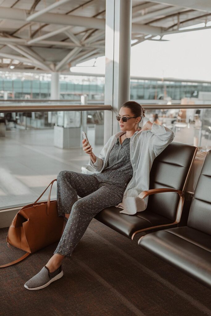 Woman in airport lounge wearing sunglasses, checking phone, seated next to brown tote bag, relaxed travel style.