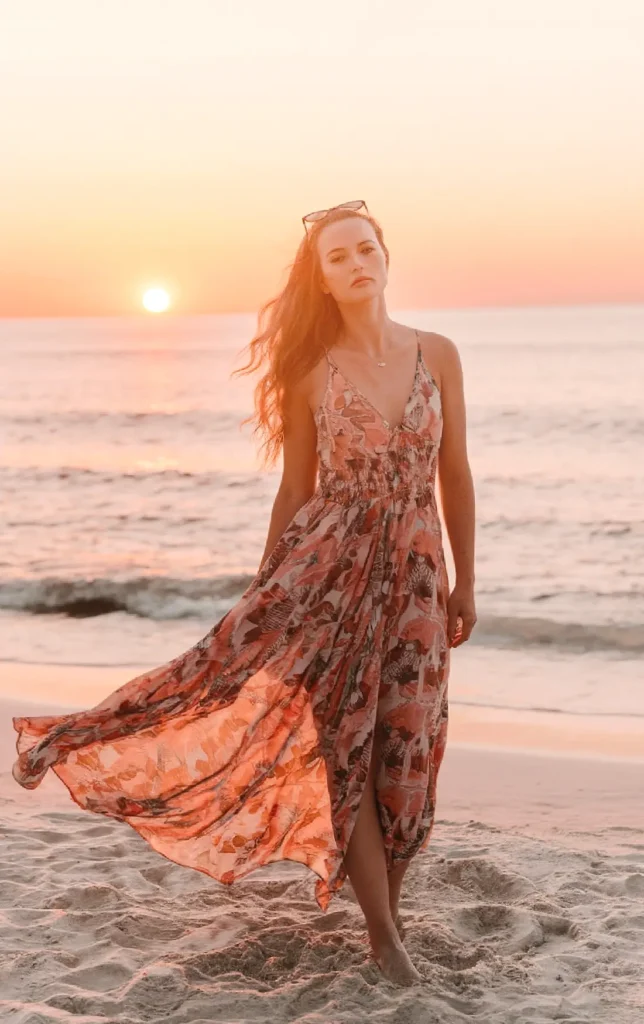 Woman in flowing dress walking on beach at sunset, hair blowing, serene ocean background.