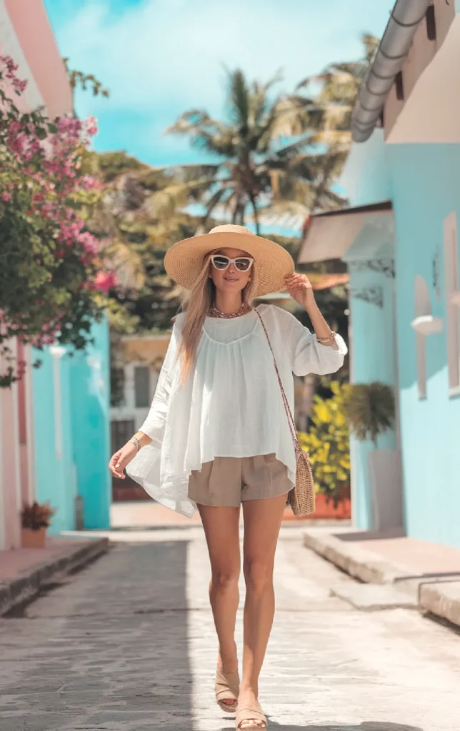 Woman in a sunhat walks down a colorful street, showcasing summer fashion with a chic white blouse and shorts.