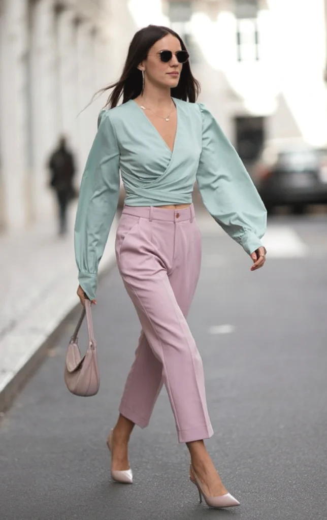 Fashionable woman in pastel outfit walking confidently, wearing sunglasses and holding a stylish bag on a city street.