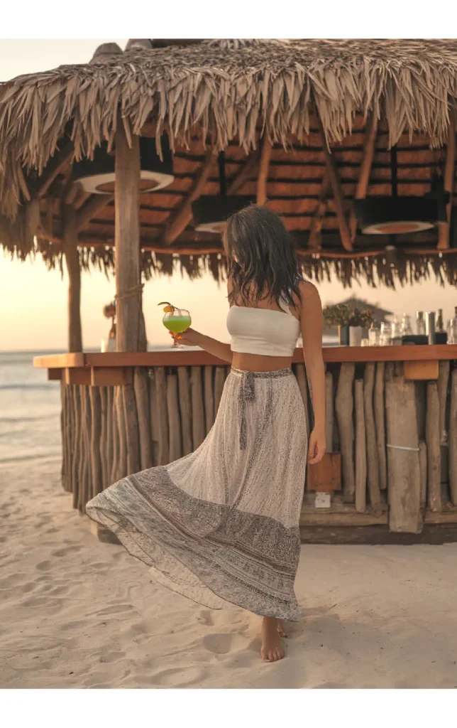 Woman in beach attire with drink by rustic tiki bar at sunset.
