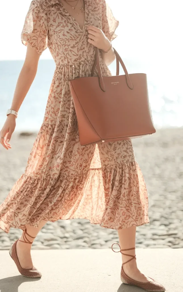 Woman in floral dress with brown tote bag walking on the beach.