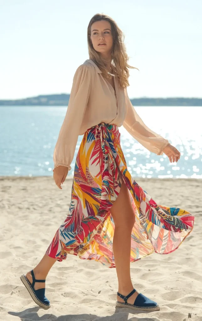 Woman in colorful skirt and blouse walking on a sunny beach, ocean in background.