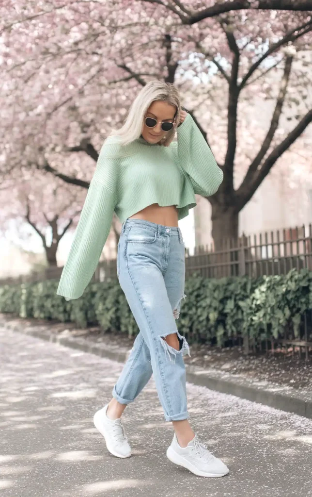 Fashionable woman in mint sweater and ripped jeans walking under cherry blossoms in spring.