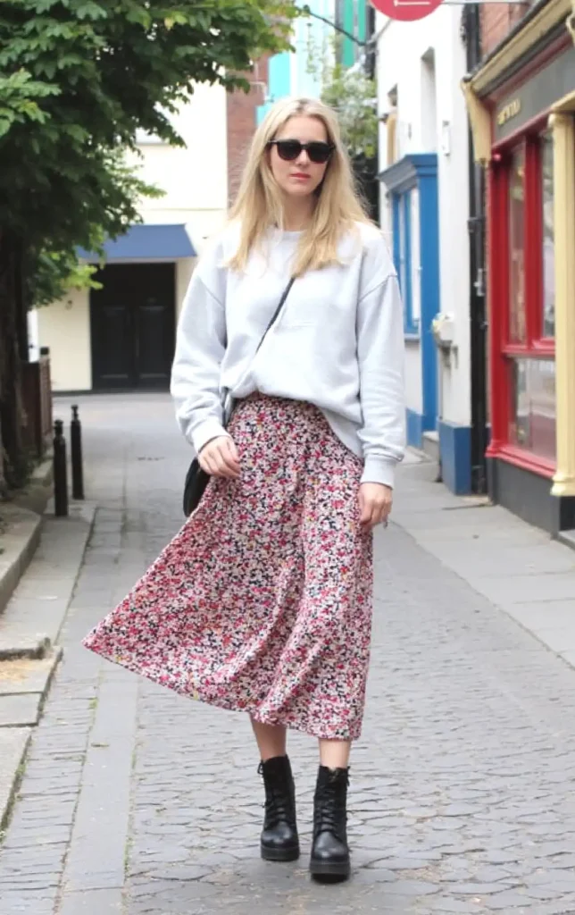 Woman in floral skirt and boots walking down a cobblestone street, wearing sunglasses and a white sweater.