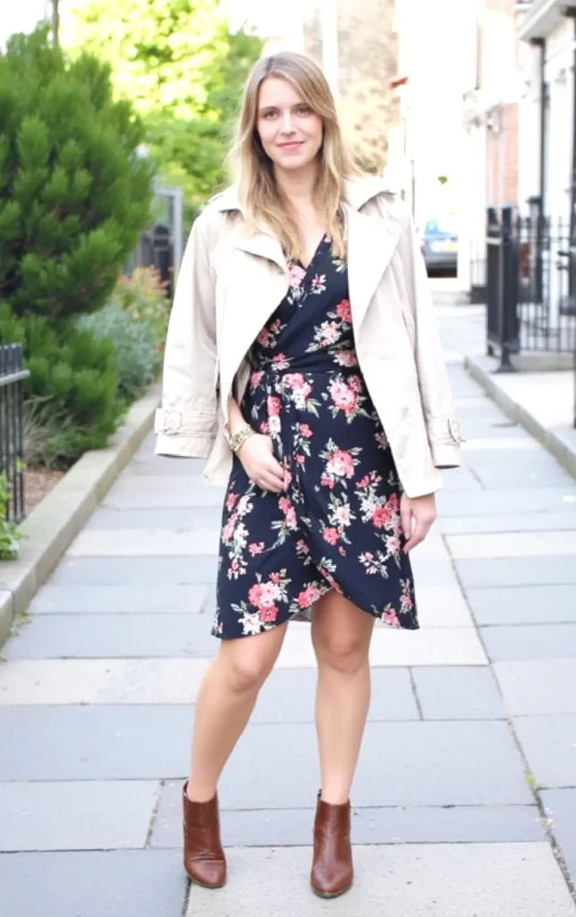 Woman in floral dress and trench coat walking on sidewalk, showcasing stylish fall fashion with ankle boots.