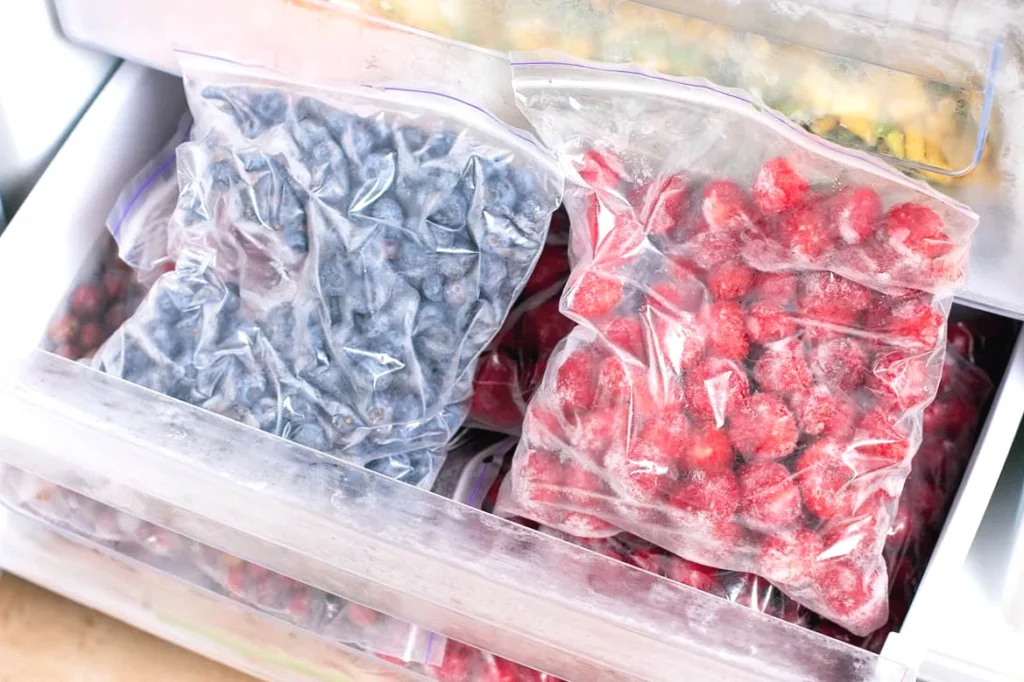 Bags of frozen blueberries and strawberries stored in a freezer drawer for fresh and healthy preservation.
