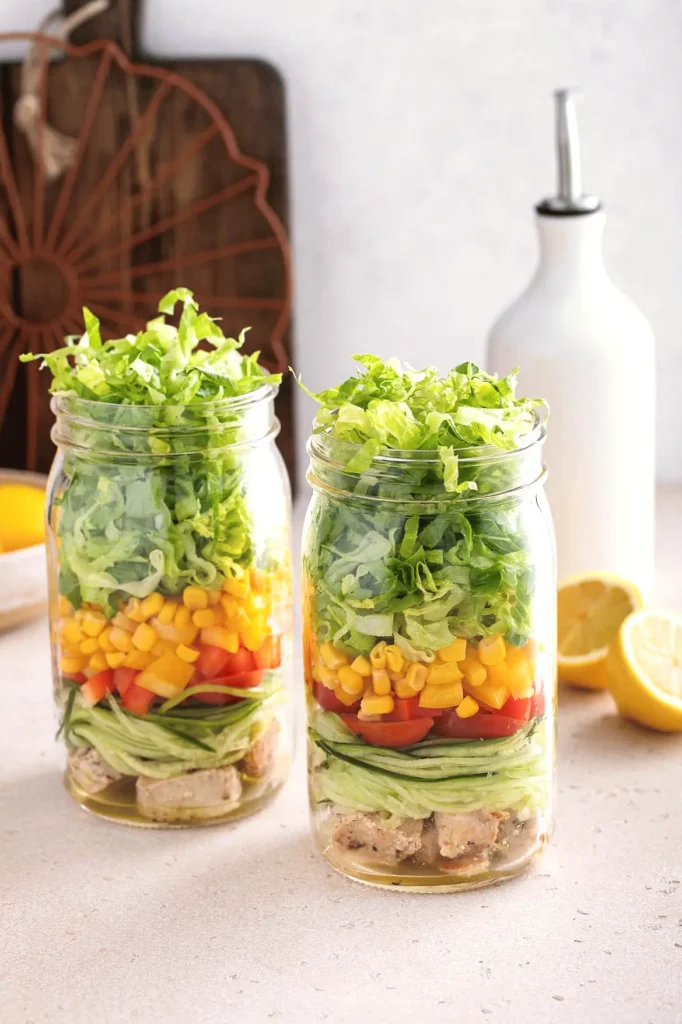 Layered salad in mason jars with lettuce, corn, peppers, tomatoes, cucumber, and chicken, next to lemon and oil bottle.