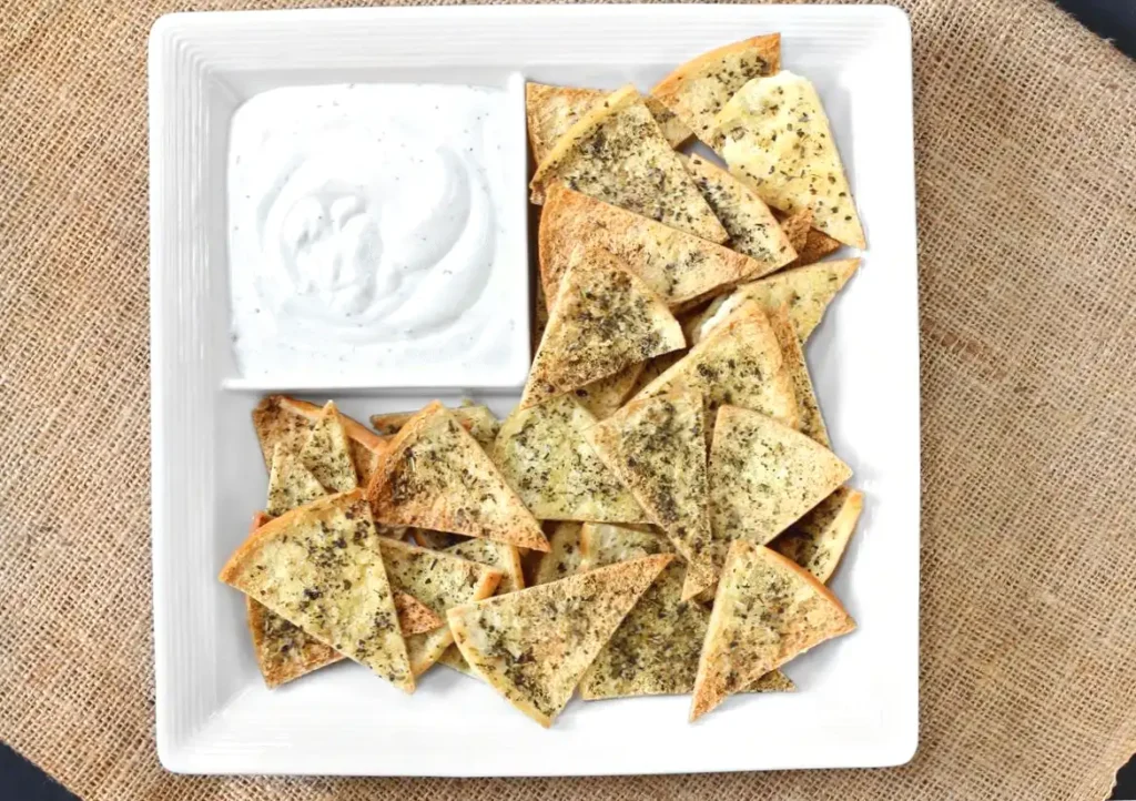 Herb-seasoned pita chips with creamy dip on a white square plate.