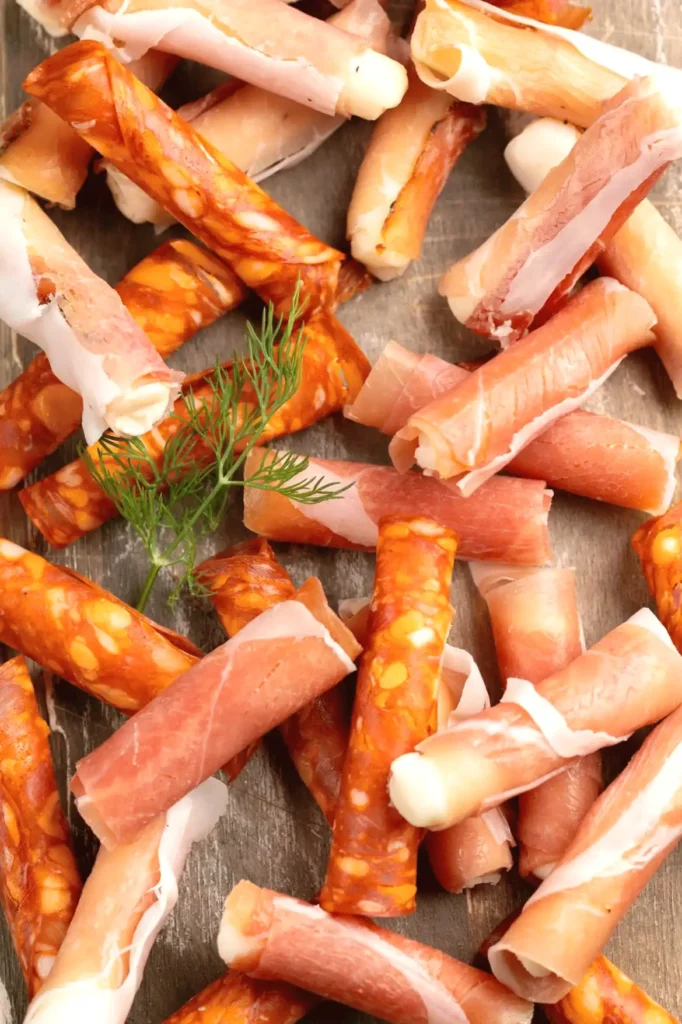 Assorted cured meat sticks with prosciutto and chorizo on a rustic wooden board, garnished with herbs.