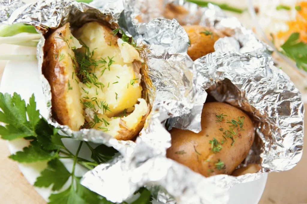 Baked potato in foil with fresh parsley and dill on a plate, showcasing a delicious, simple, homemade meal.