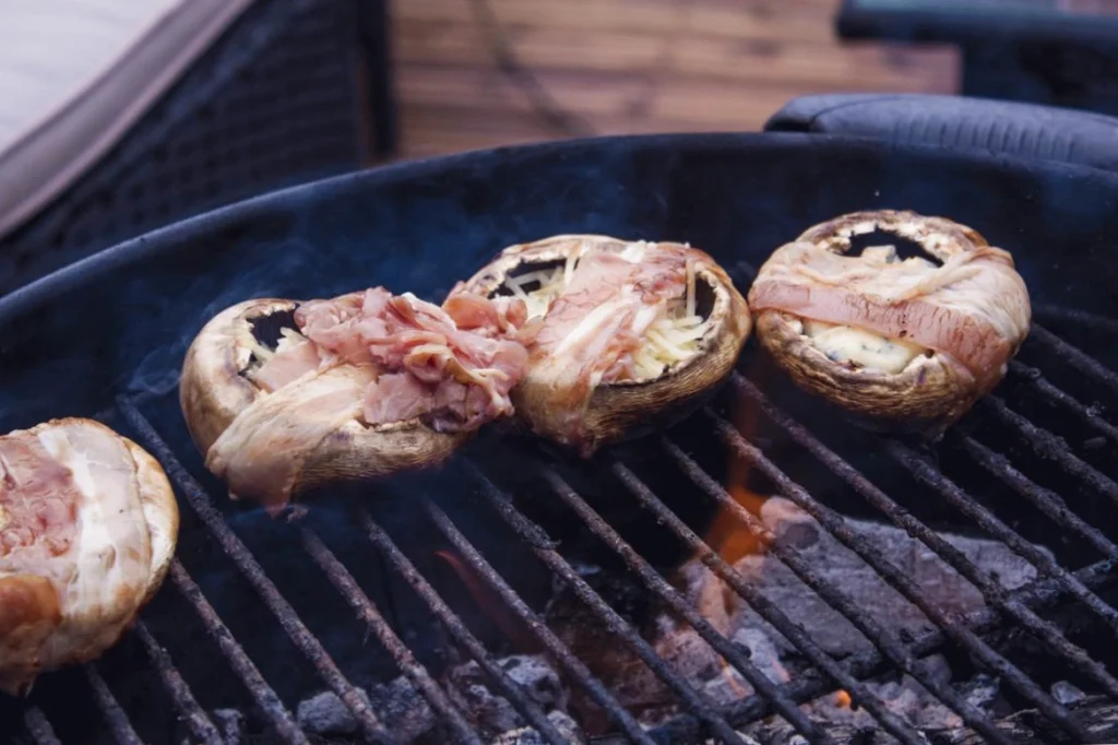 Grilled mushrooms stuffed with bacon and cheese, sizzling on outdoor barbecue grill.