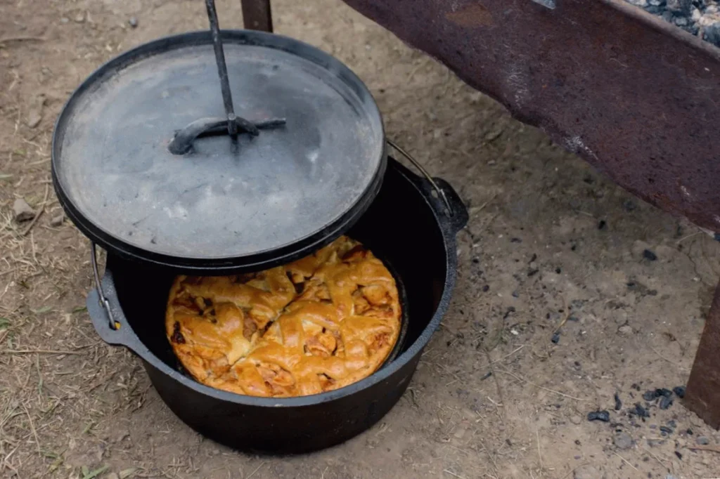 Dutch oven with a freshly baked pie, partially open lid, placed on outdoor ground near a fire pit.