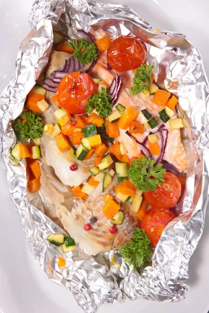 foil packets for camping, grilled fish in foil with cherry tomatoes, zucchini, carrots, and parsley garnish. Healthy and colorful meal.