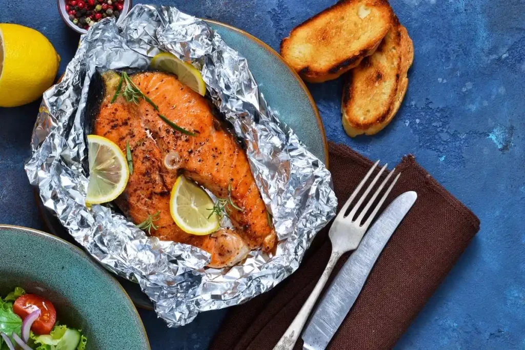 Grilled salmon with lemon slices on foil, served with toasted bread and fresh salad on a blue table setting.