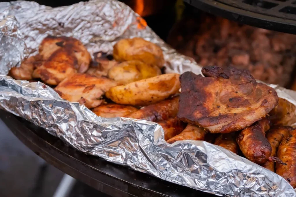 Grilled meats sizzling on foil, showcasing crispy chicken and sausages at a barbecue. Perfect for outdoor grilling enthusiasts.