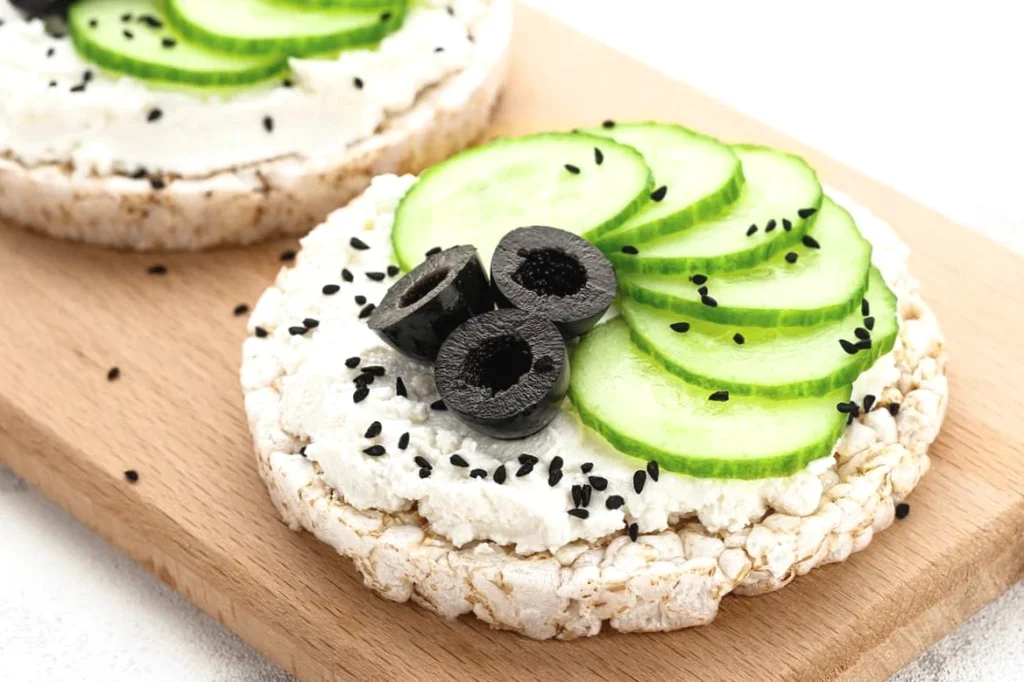Rice cake topped with cheese, cucumber slices, black olives, and nigella seeds on a wooden board.