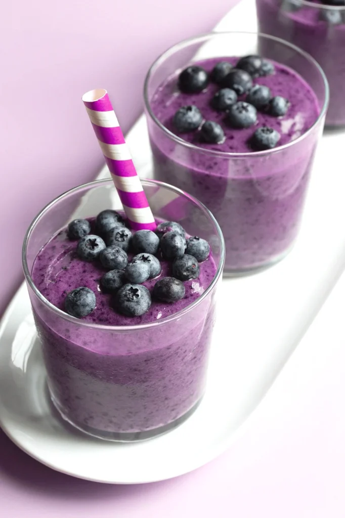 Purple blueberry smoothie in glass with striped straw on a plate, topped with fresh blueberries.