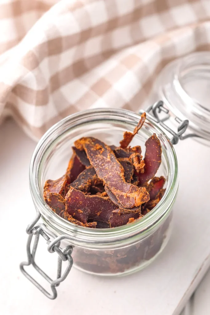 Glass jar filled with spiced biltong slices, perfect protein snack, on checkered cloth background.