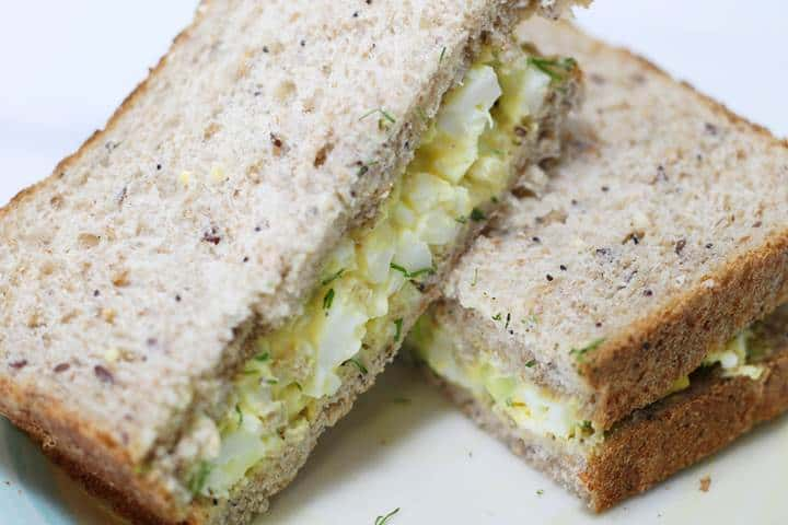 Egg salad sandwich on whole grain bread, sliced and ready to eat on a white plate, garnished with herbs.