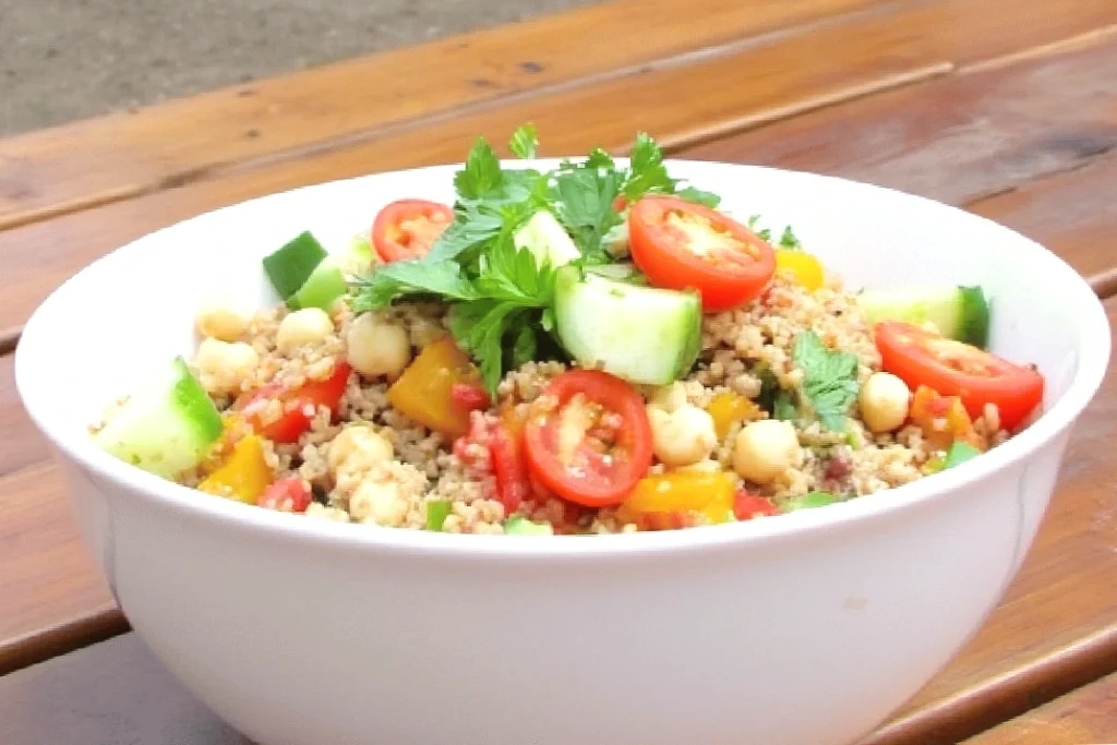 Fresh Mediterranean couscous salad with cherry tomatoes, cucumber, and chickpeas in a white bowl on a wooden table.