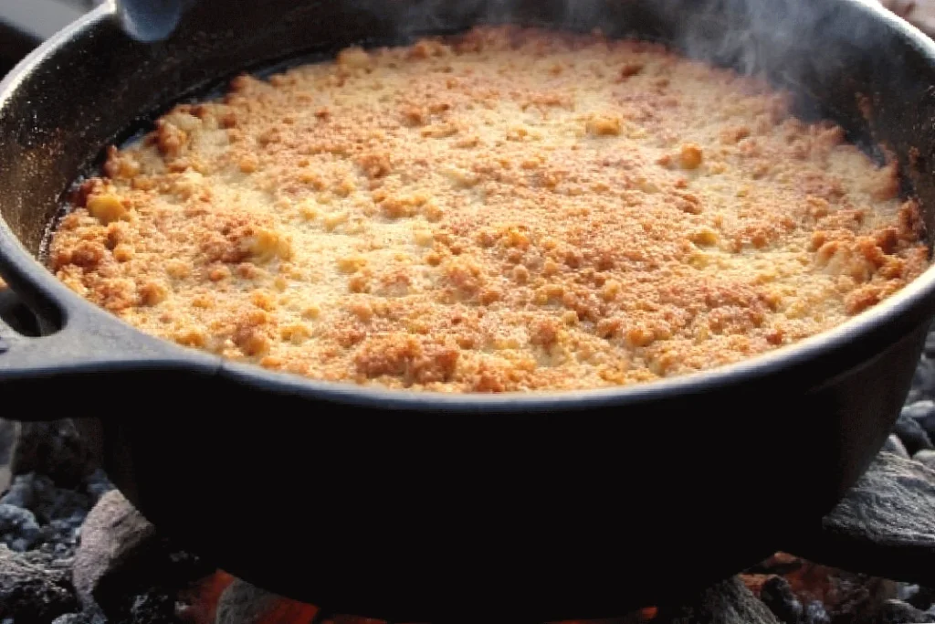 Baked apple cobblers with a golden crust in a black cast-iron skillet, cooked over an outdoor fire.