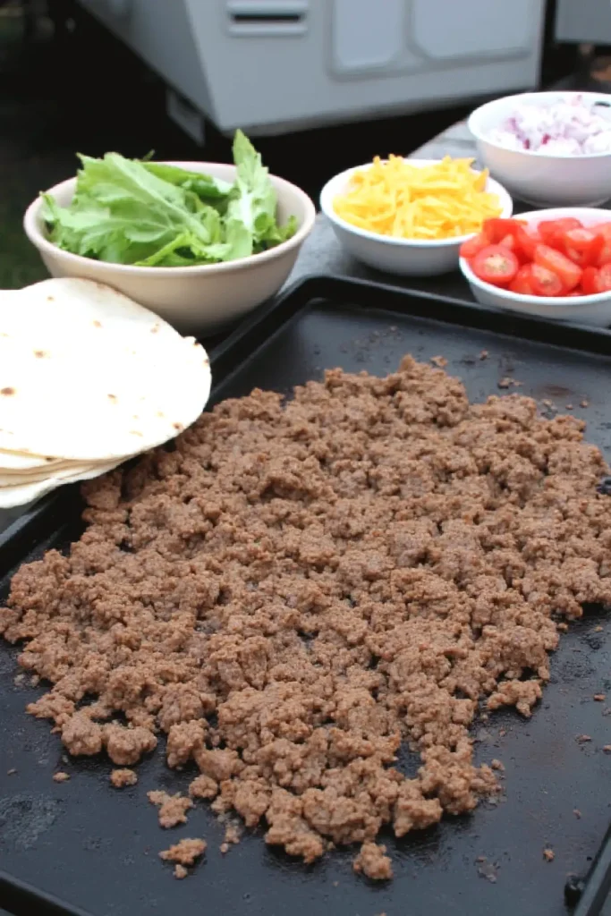 Ground beef, tortillas, lettuce, cheese, tomatoes, and onions on a grill for tacos. Perfect for a delicious taco meal.