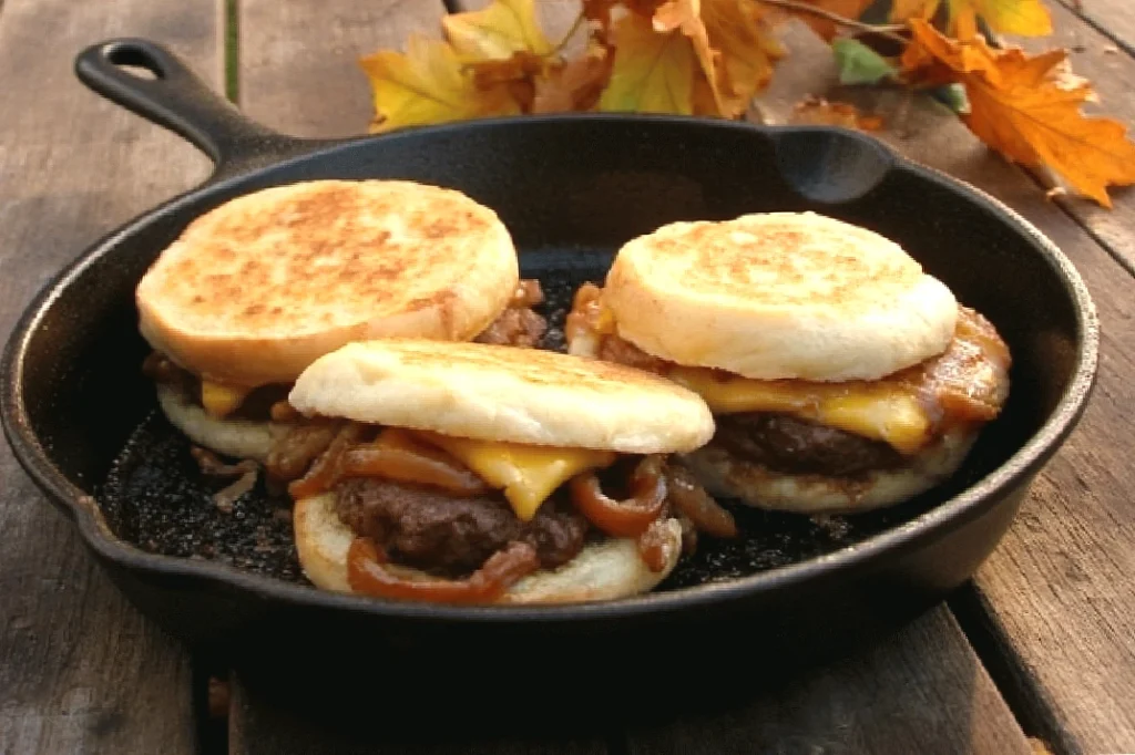 Three juicy burgers with cheese and onions in a skillet, set against autumn leaves on a wooden table.