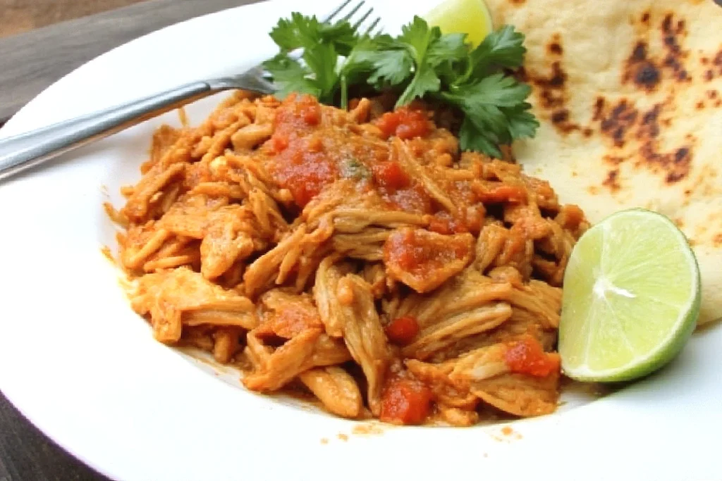 Shredded chicken tinga with tomato sauce, lime wedge, and cilantro on a plate with a tortilla.