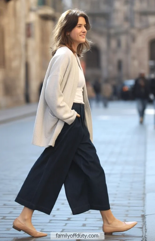 Woman walking in city, wearing beige sweater, white top, and wide-leg pants. Casual chic street fashion.