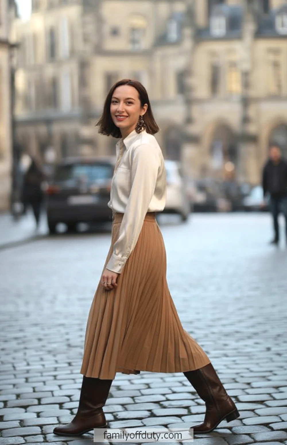 Woman walking in a stylish beige skirt and boots on a cobblestone street, elegant European urban setting.
