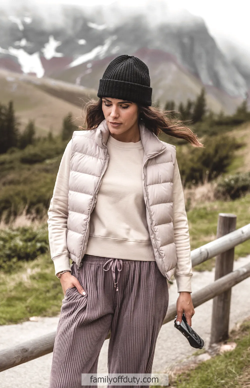 Mountain casual outfit ideas. Woman in winter attire walking on a mountain path, wearing a beanie and puffer vest.