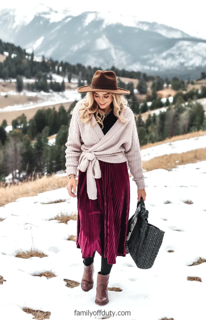Woman in winter outfit walking in snowy mountains, wearing a brown hat and red skirt, smiling.