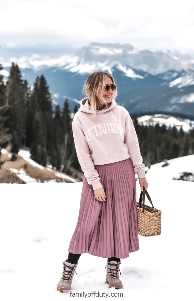 Woman in a pink hoodie and skirt standing in snowy mountains with a basket, wearing sunglasses.