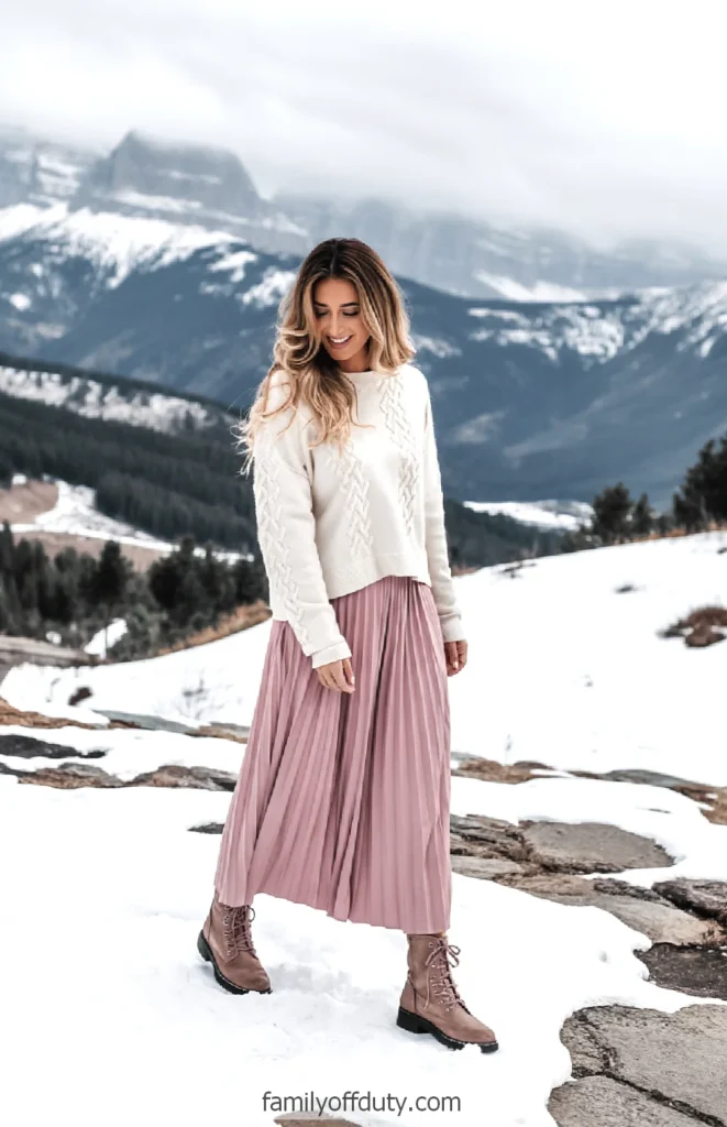 Woman in a white sweater and pink skirt walking in snow-covered mountains, smiling warmly amidst stunning scenery.