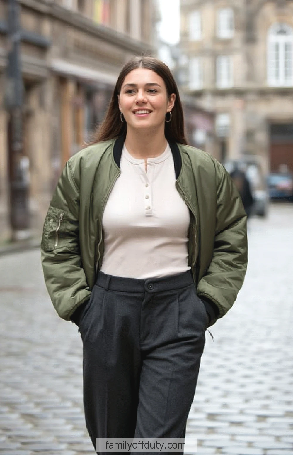 Europe winter outfit ideas. Smiling woman in green jacket and white shirt walks on cobblestone street, urban background.
