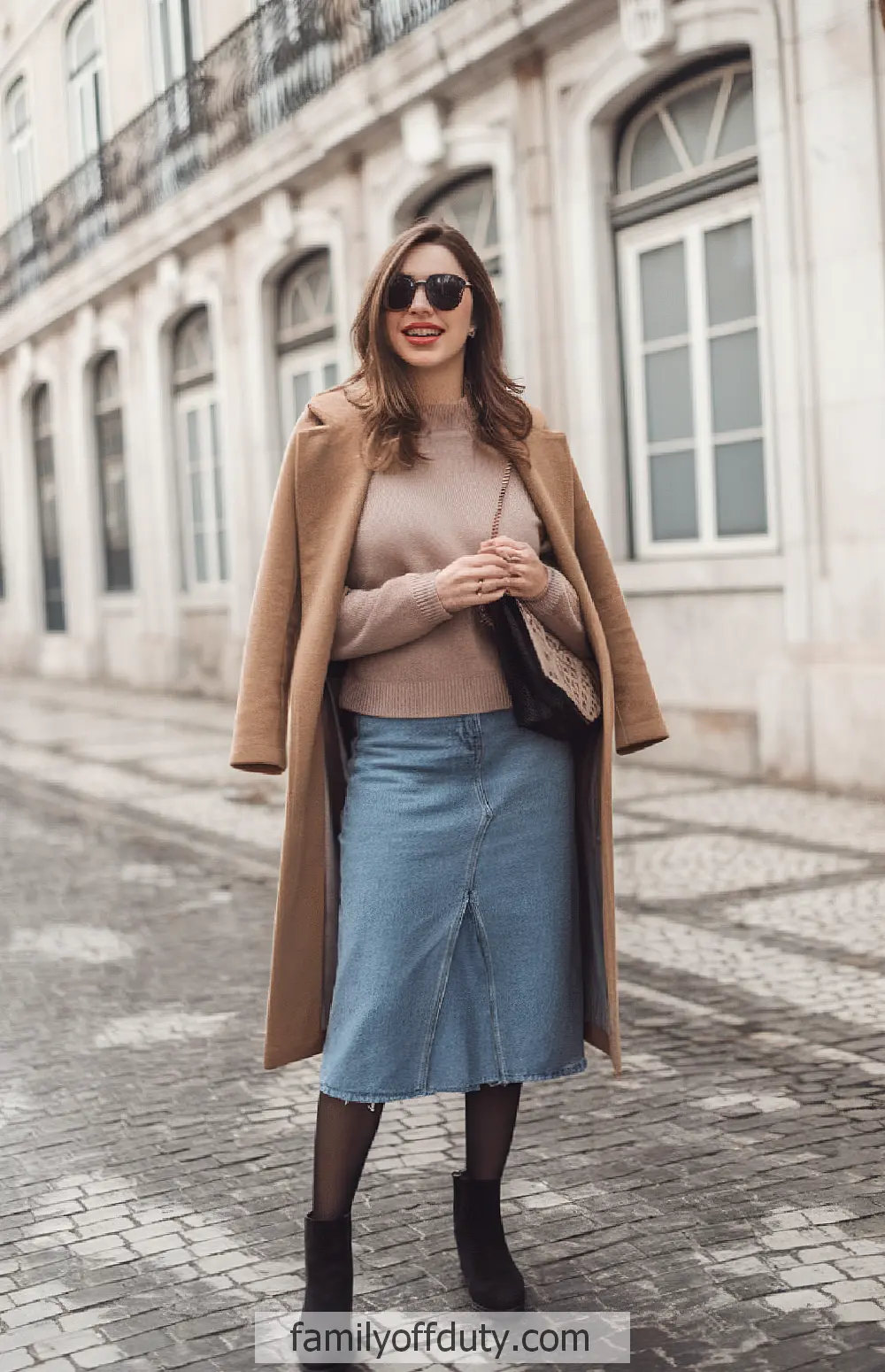 Fashionable woman in denim skirt and camel coat smiling on cobblestone street.