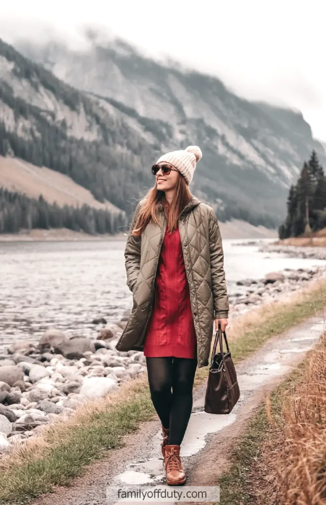 Woman in winter coat and hat walking by a scenic river with mountains in the background.