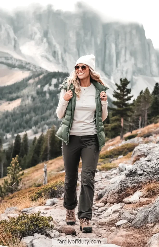 Woman hiking in scenic mountain landscape wearing a green vest and white beanie, enjoying the outdoors.