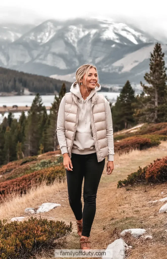 Smiling woman in a puffer vest hiking in a scenic mountain landscape with cloudy skies.