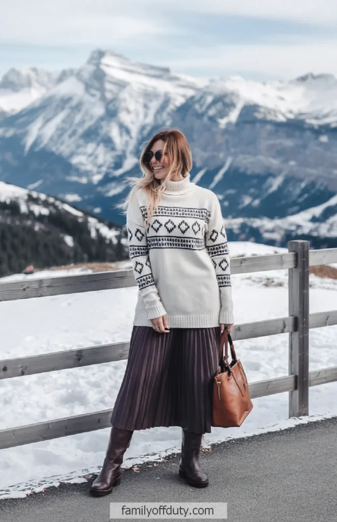 Woman in winter fashion with snowy mountain background, wearing sweater and pleated skirt, holding a leather bag.