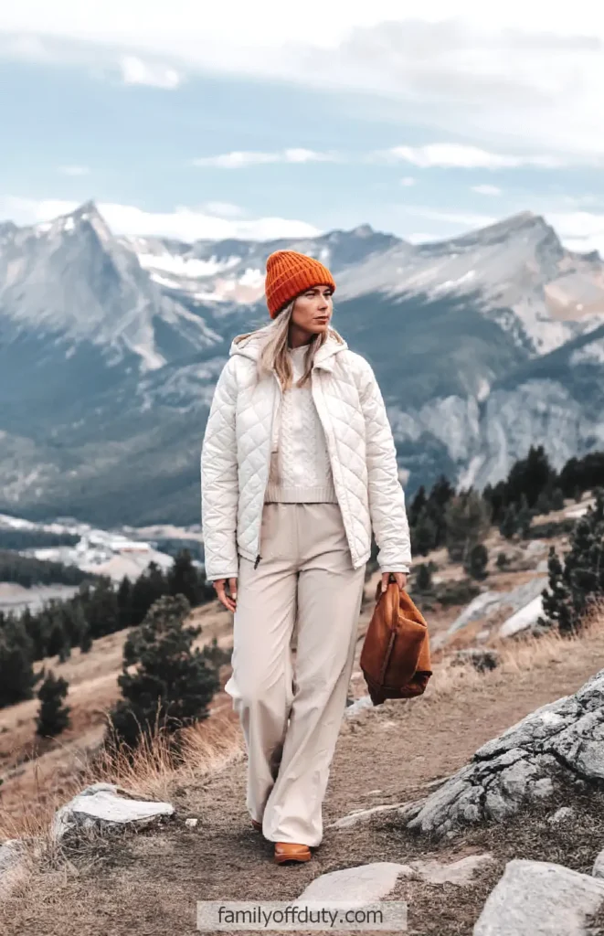 Woman in winter hiking attire and orange beanie walking through scenic mountain landscape.