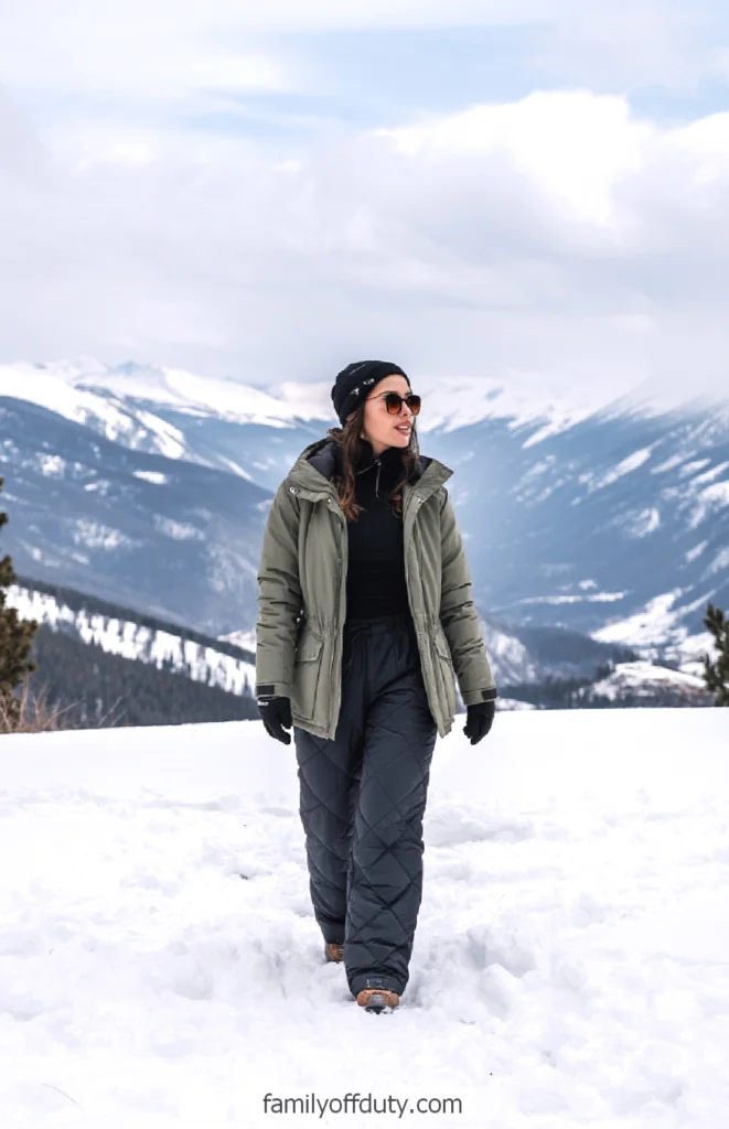 Woman in winter clothing walking in snowy mountain landscape with trees in background.